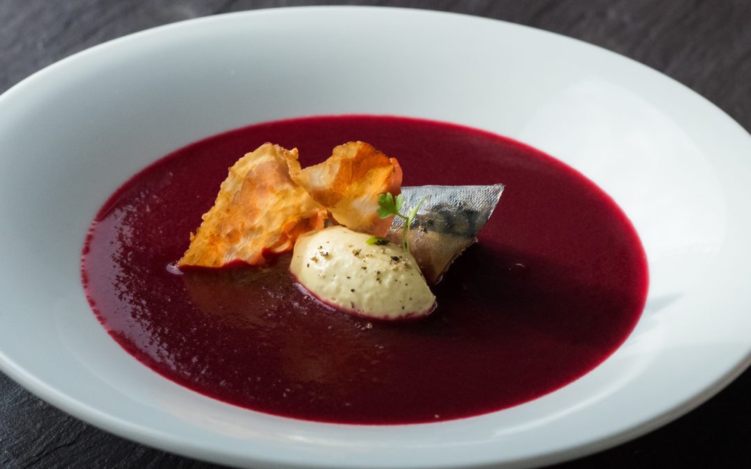 A white bowl filled with deep claret coloured soup, with a central garnish of smoked mackarel, horseradish cream and crispy pancetta