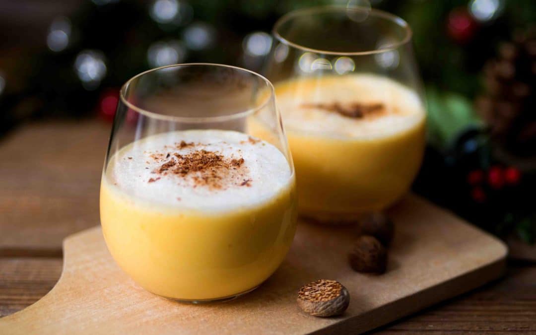 Two glasses of golden yellow eggnong in a warm, twinkling Christmas scene w