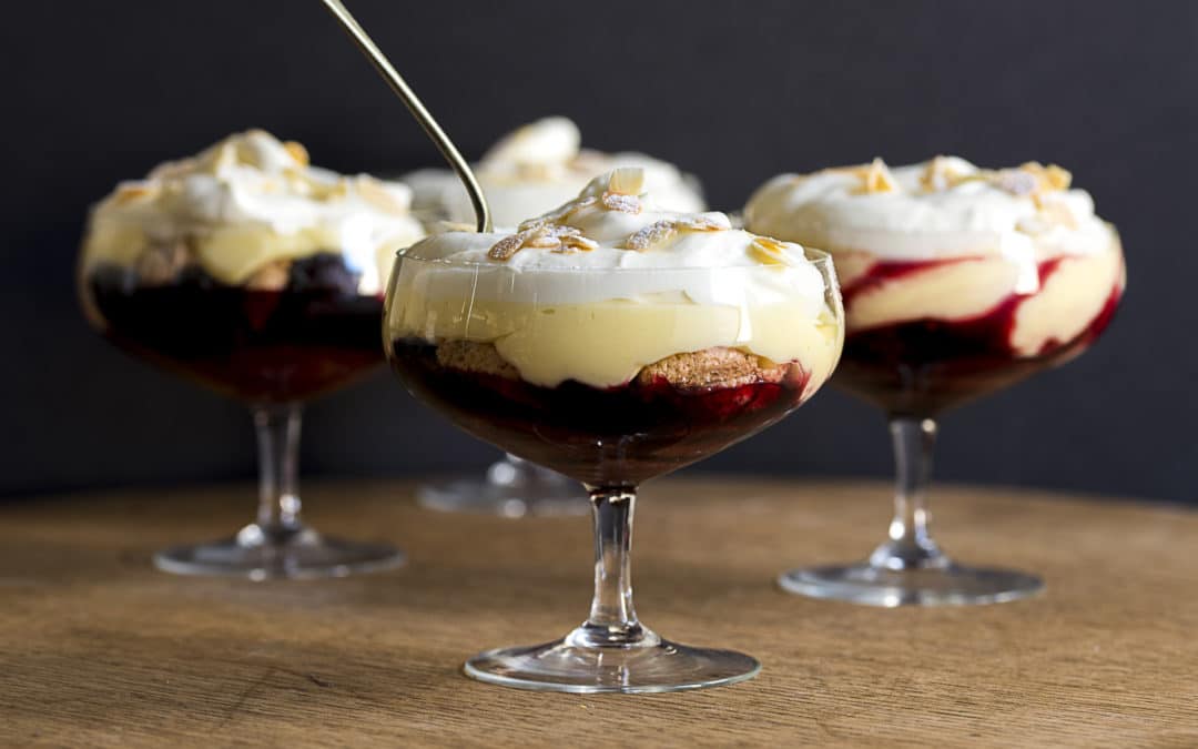 a glass showing layers of blackberry and amaretto trifle, custard and topped with cream and flaked almonds